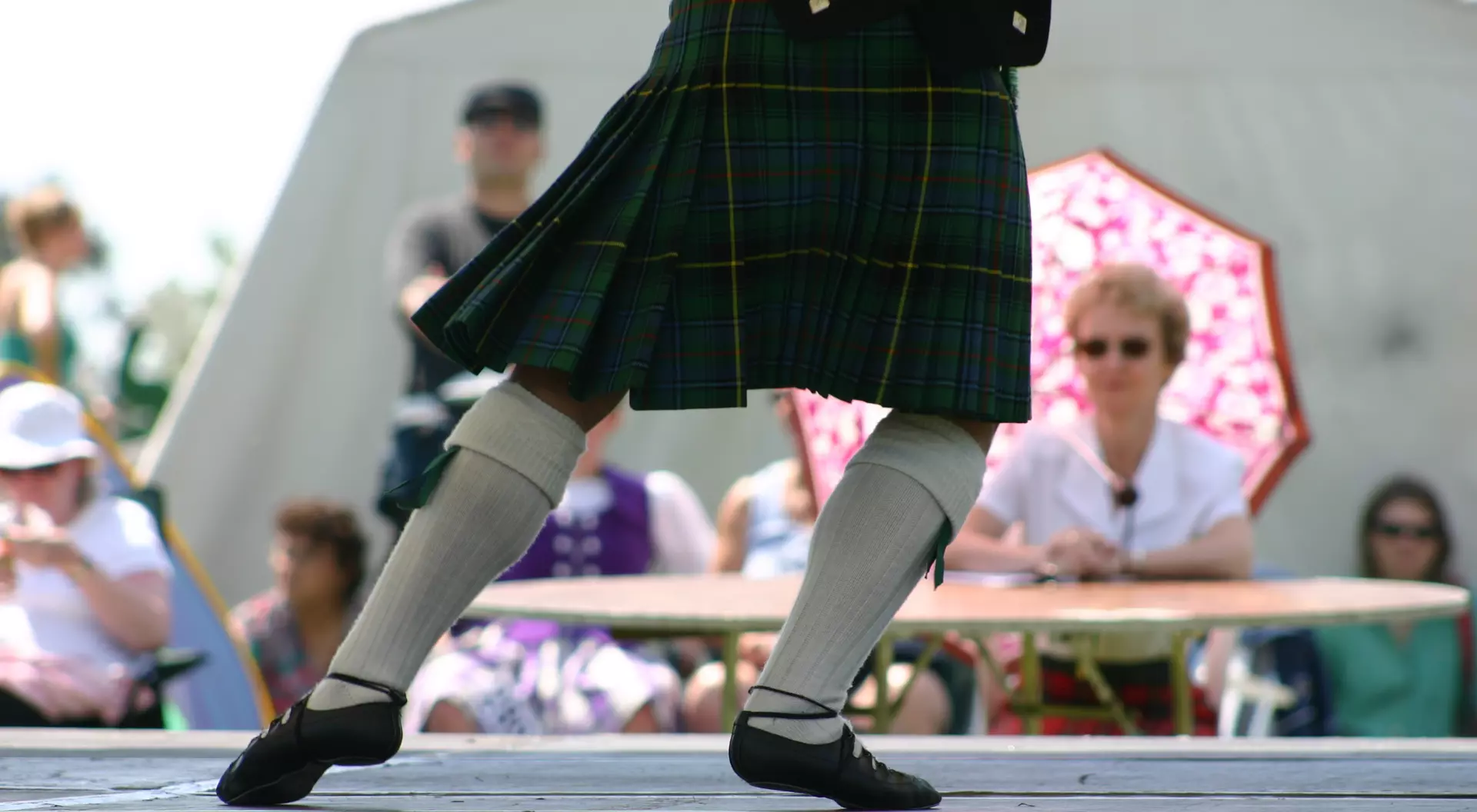 A dancer in a kilt performs on stage, while an audience watches, some under a colorful umbrella.