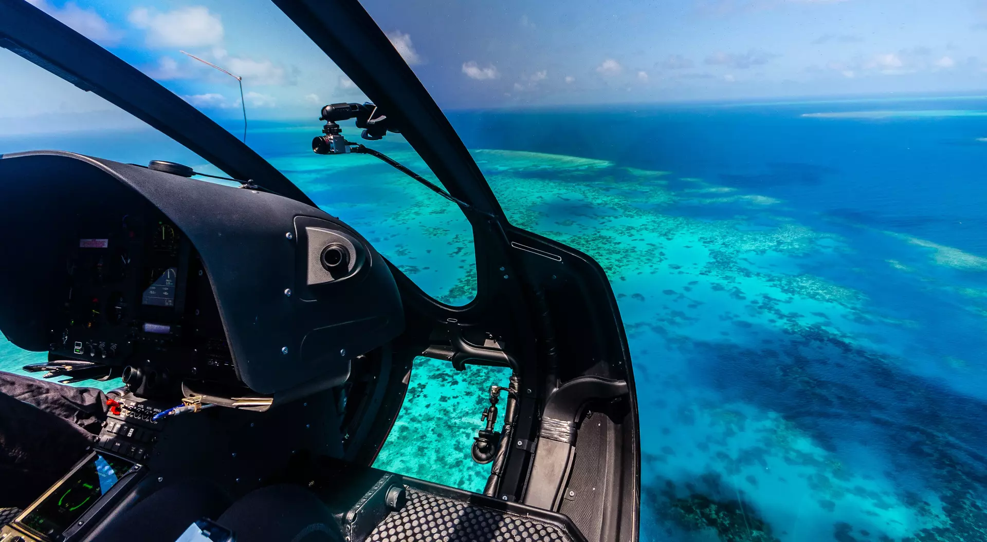 Cockpit view of a helicopter flying over vibrant turquoise waters and coral reefs.