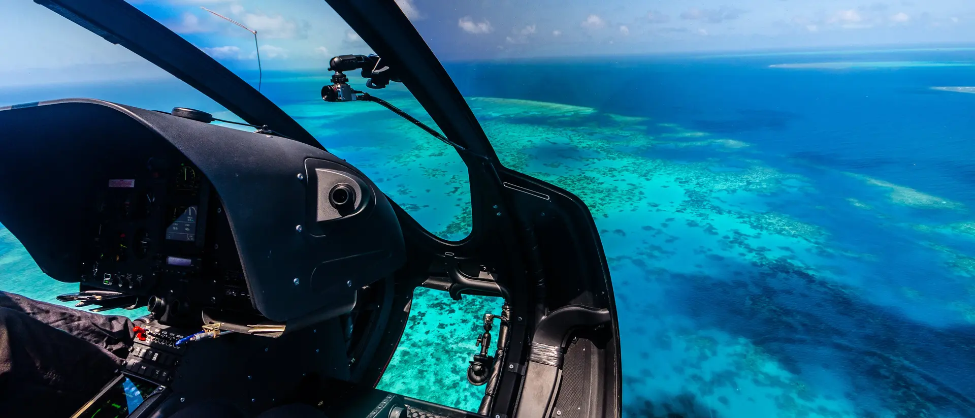 Cockpit view of a helicopter flying over vibrant turquoise waters and coral reefs.