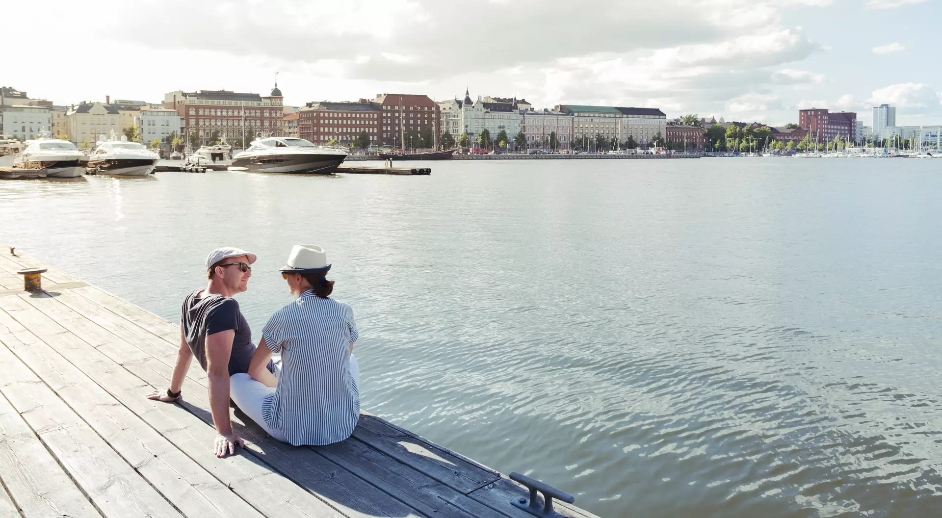 Two people sitting on a dock by the water, with boats in the background and city buildings along the shore.