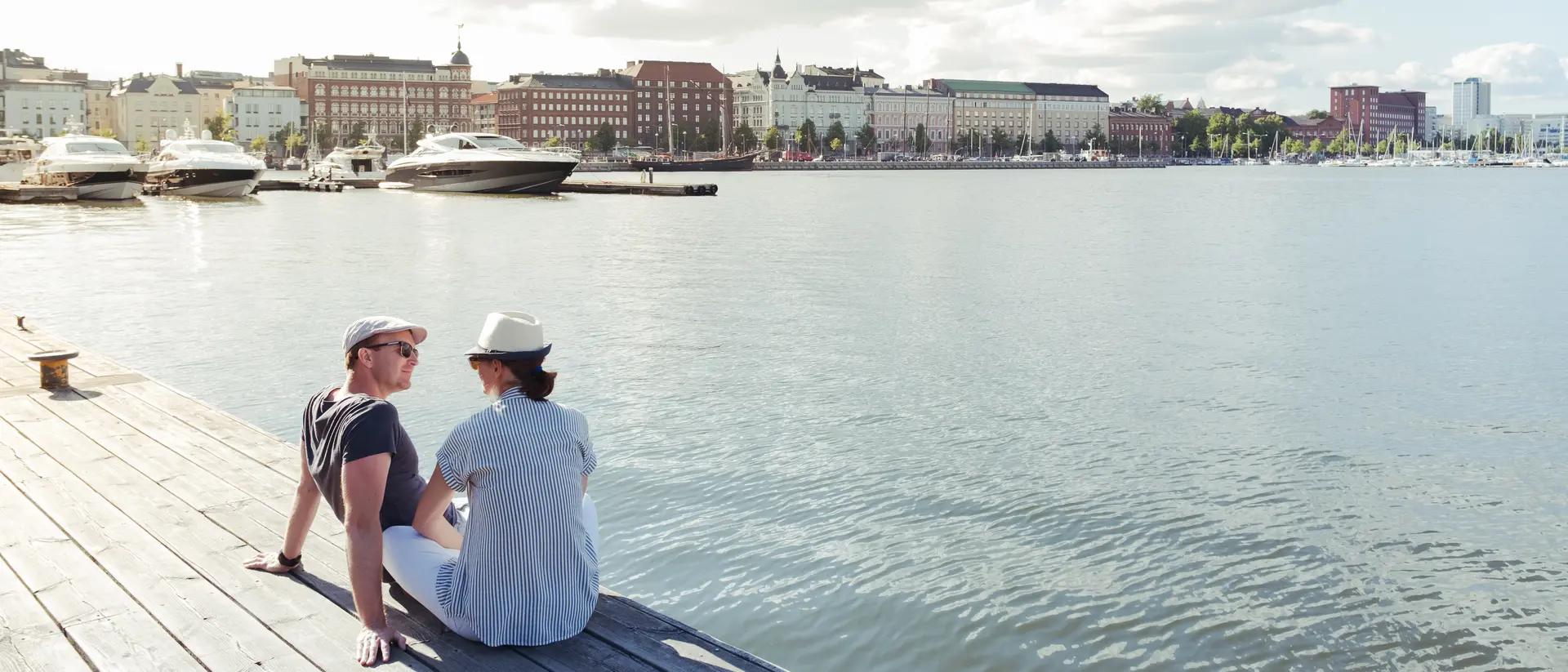 Two people sitting on a dock by the water, with boats in the background and city buildings along the shore.