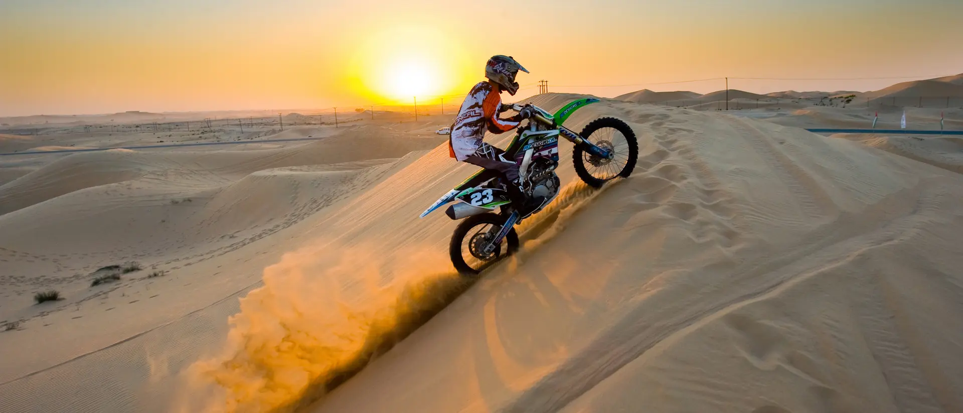 A motocross rider ascends a sand dune at sunset, kicking up dust in a dramatic desert landscape.