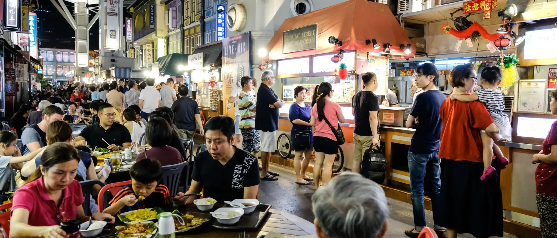 Busy night market scene with people dining and standing near food stalls, surrounded by colorful shopfronts.