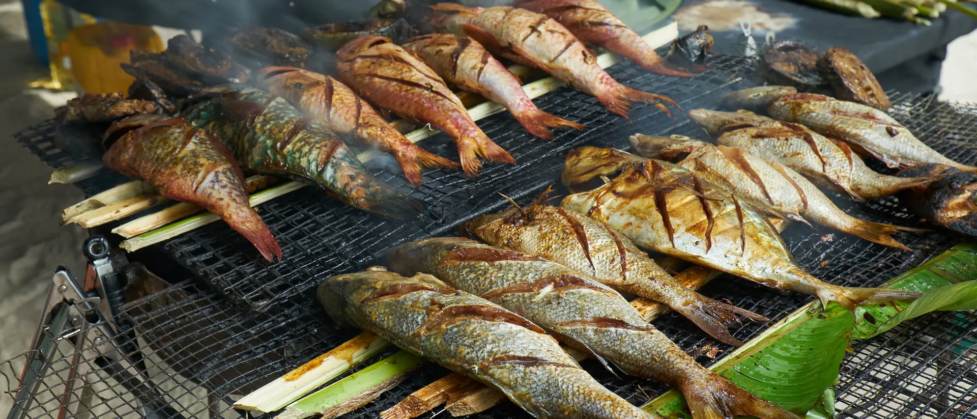 Grilled fish on a charcoal grill, with smoke rising, surrounded by green leaves and a beach setting in the background.