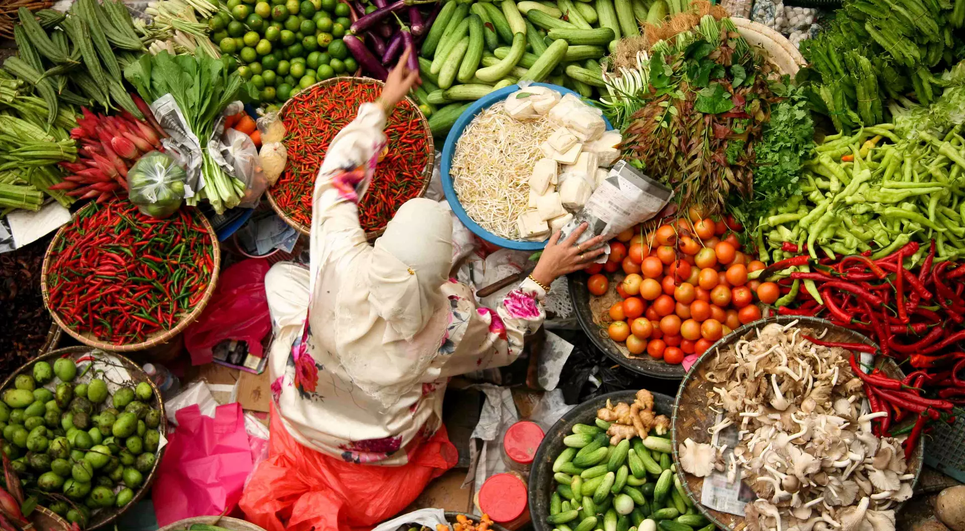 A vendor in a market reaches for produce among vibrant displays of fruits and vegetables in various baskets.