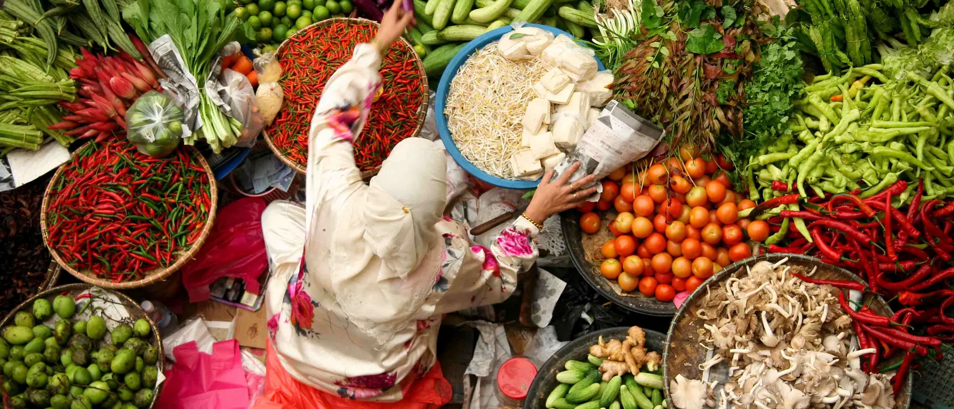 A vendor in a market reaches for produce among vibrant displays of fruits and vegetables in various baskets.
