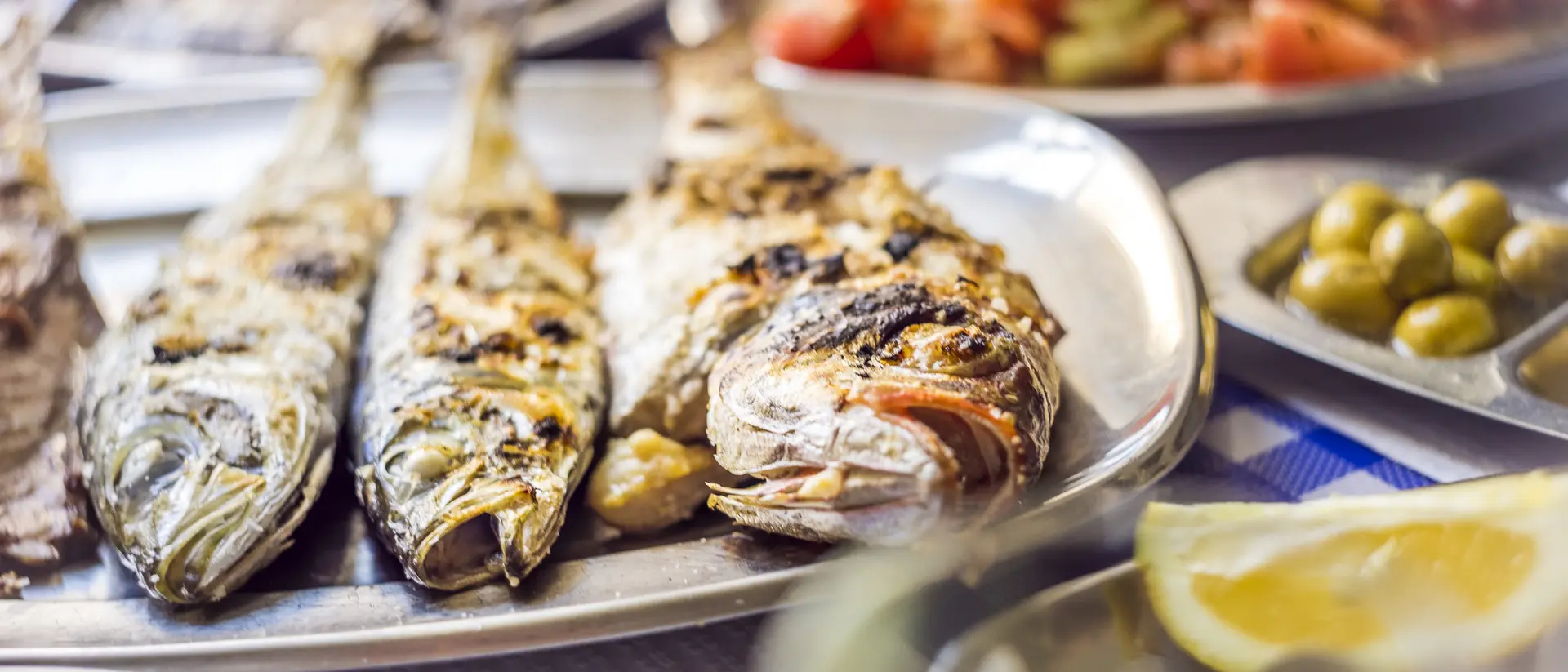 Grilled fish on a metal platter, with side dishes of olives, lemon, and mixed vegetables in the background.