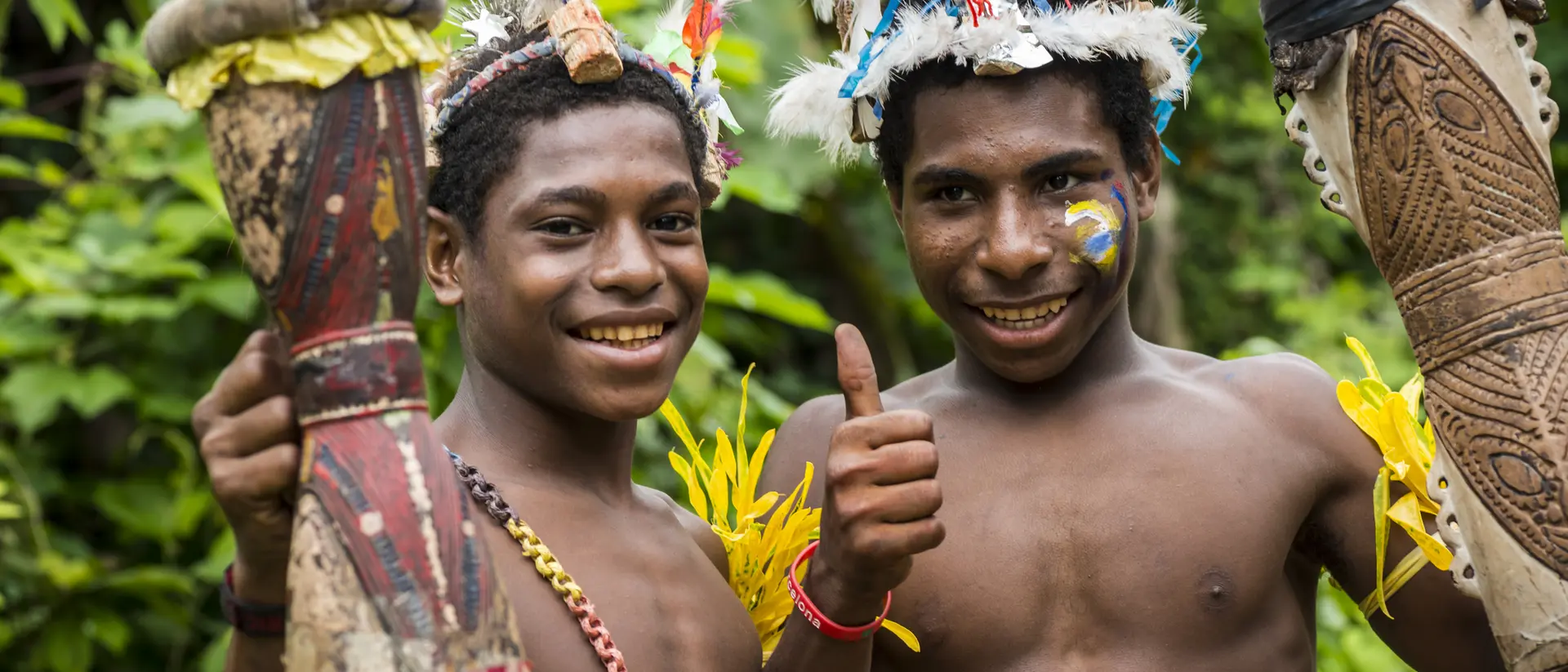 Two smiling young boys in tribal attire hold drums and give a thumbs up, surrounded by lush greenery.