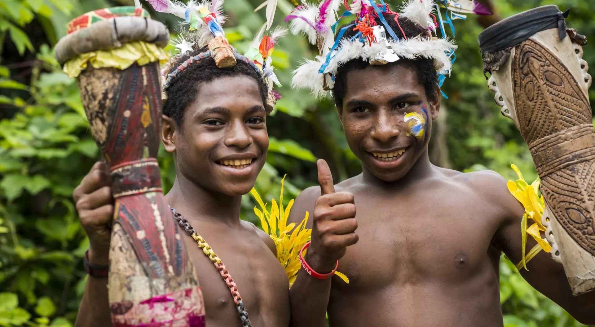 Two smiling young boys in tribal attire hold drums and give a thumbs up, surrounded by lush greenery.