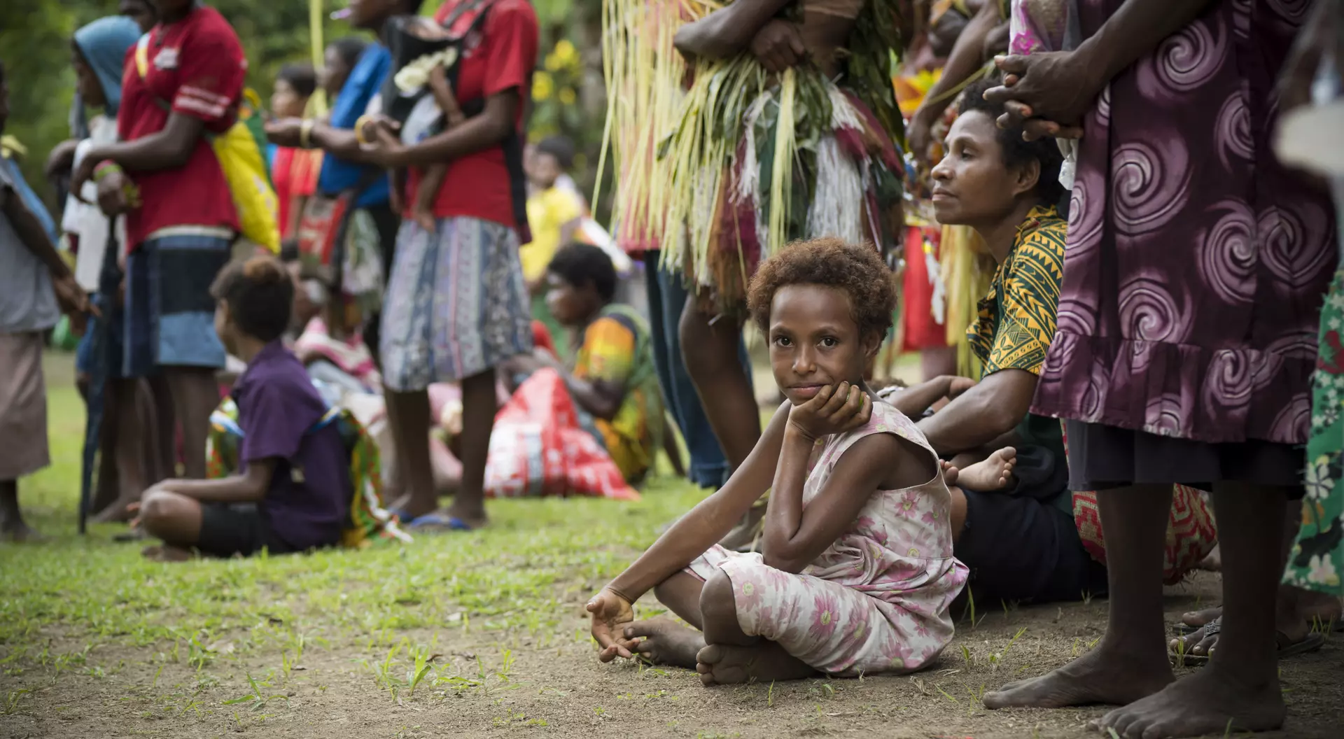 A group of people gather outdoors; a child sits in the foreground, looking contemplative amidst the crowd.