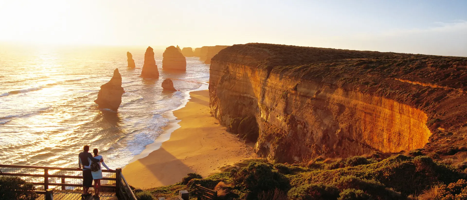 Couple standing on a wooden platform overlooking a beach with towering rock formations at sunset.