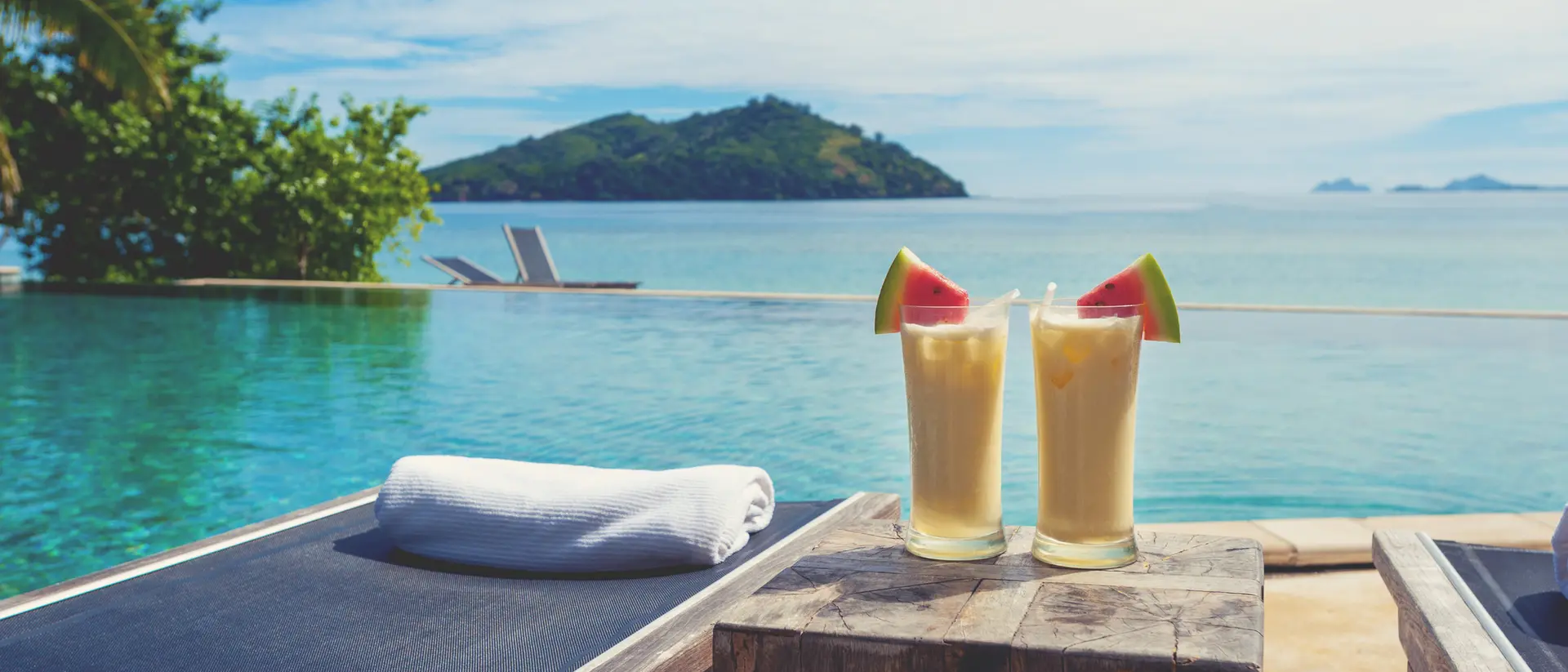Two tropical cocktails sit on a wooden table by a serene infinity pool, with lush greenery and mountains in the background.