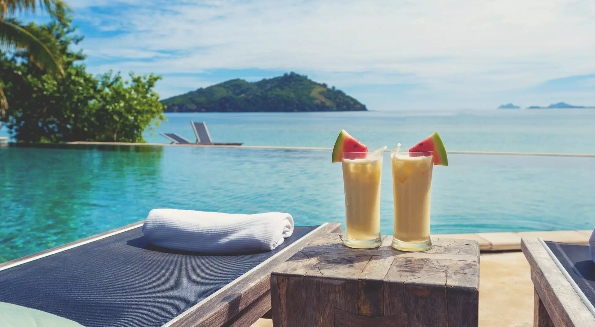 Two tropical cocktails sit on a wooden table by a serene infinity pool, with lush greenery and mountains in the background.