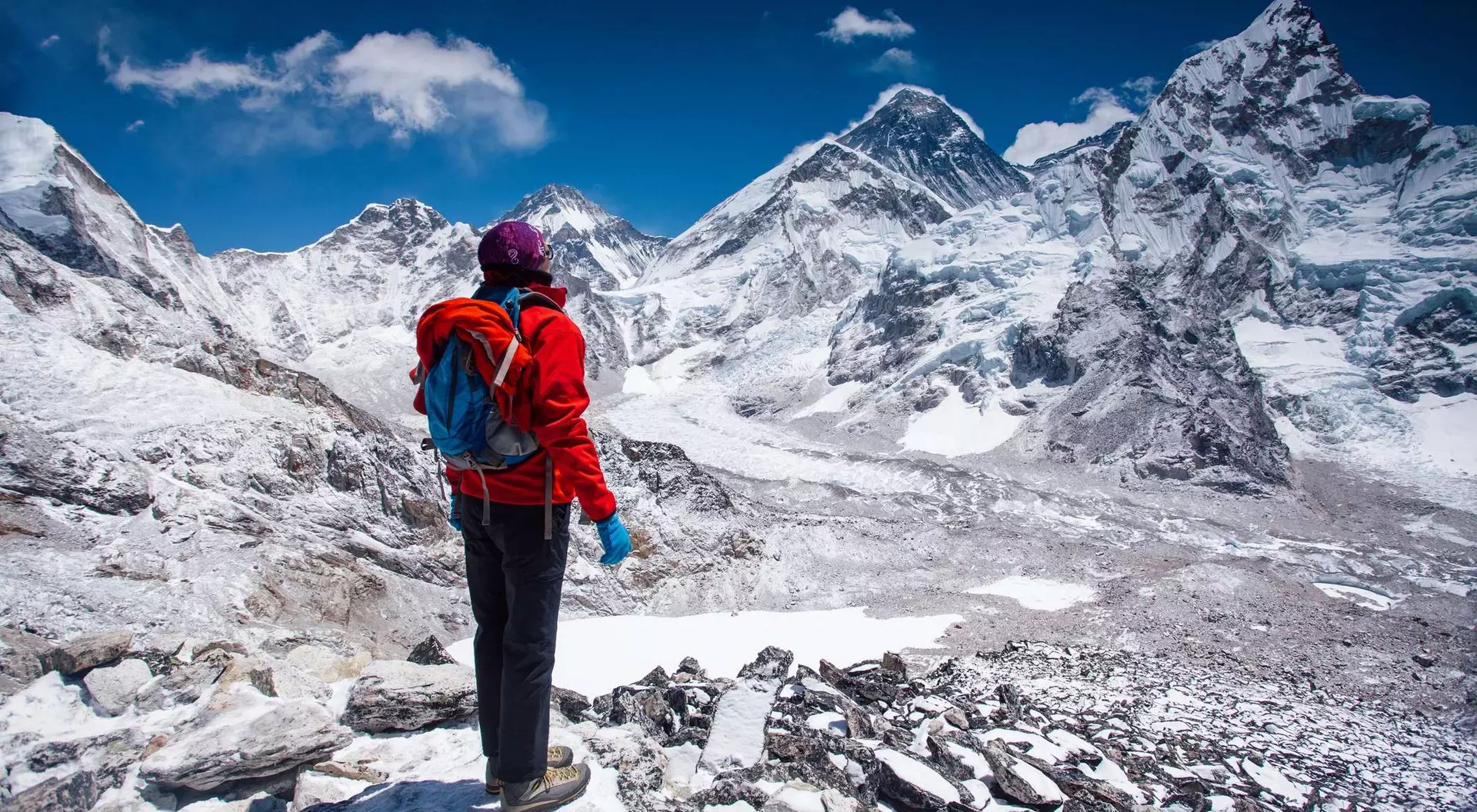 Person in a red jacket stands on rocky terrain, overlooking snow-capped mountains under a clear blue sky.