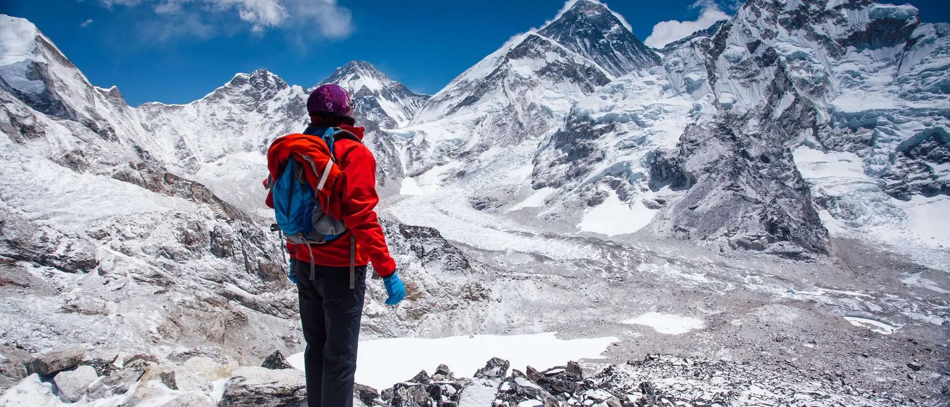 Person in a red jacket stands on rocky terrain, overlooking snow-capped mountains under a clear blue sky.