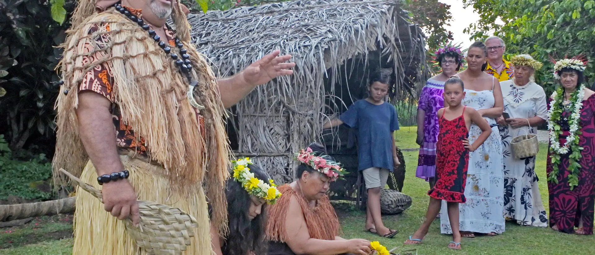 A group of people, including a man in traditional attire, gather near a thatched hut for a cultural presentation.