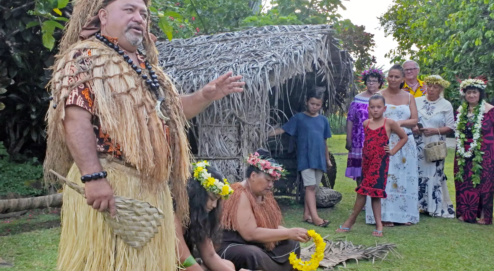 A group of people, including a man in traditional attire, gather near a thatched hut for a cultural presentation.