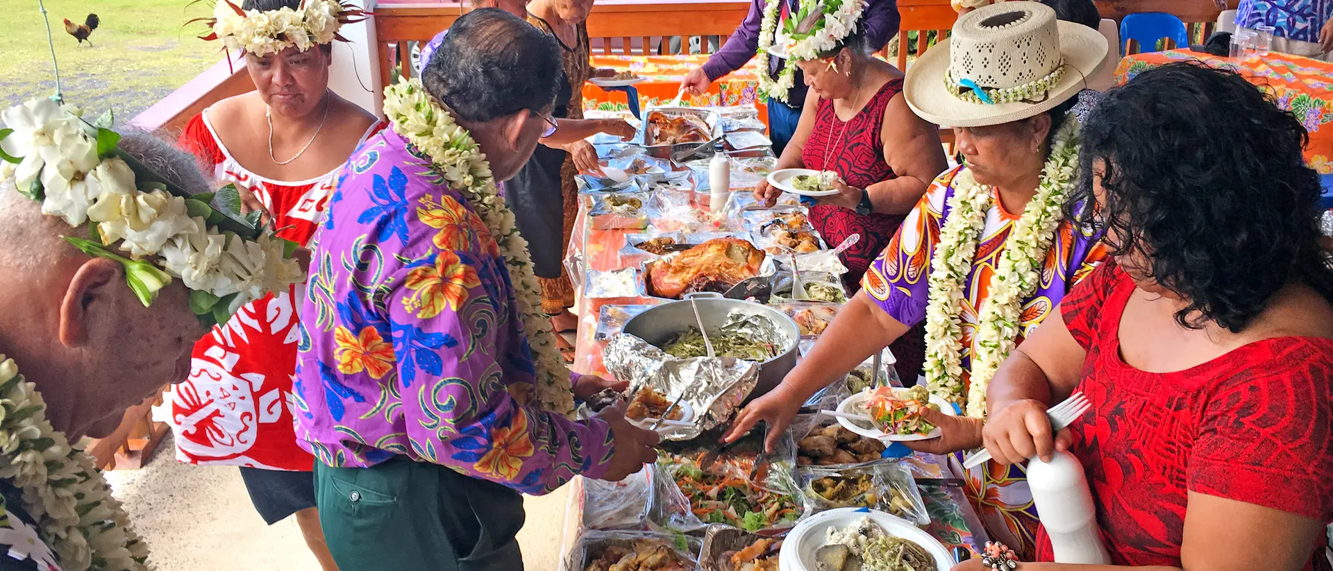 A group of people in colorful attire share a buffet spread outdoors, adorned with leis, enjoying a festive meal together.
