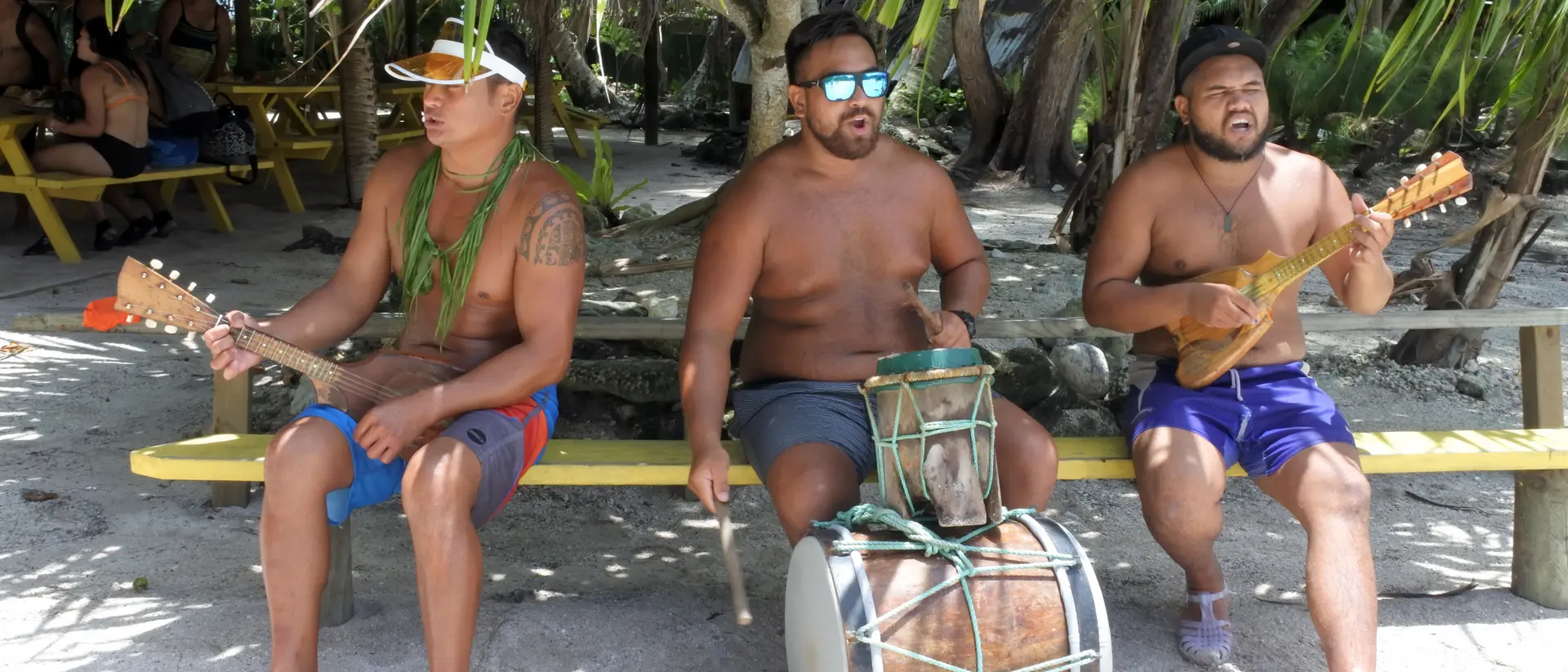 Three shirtless men play traditional instruments, with palm trees and a sandy beach in the background.