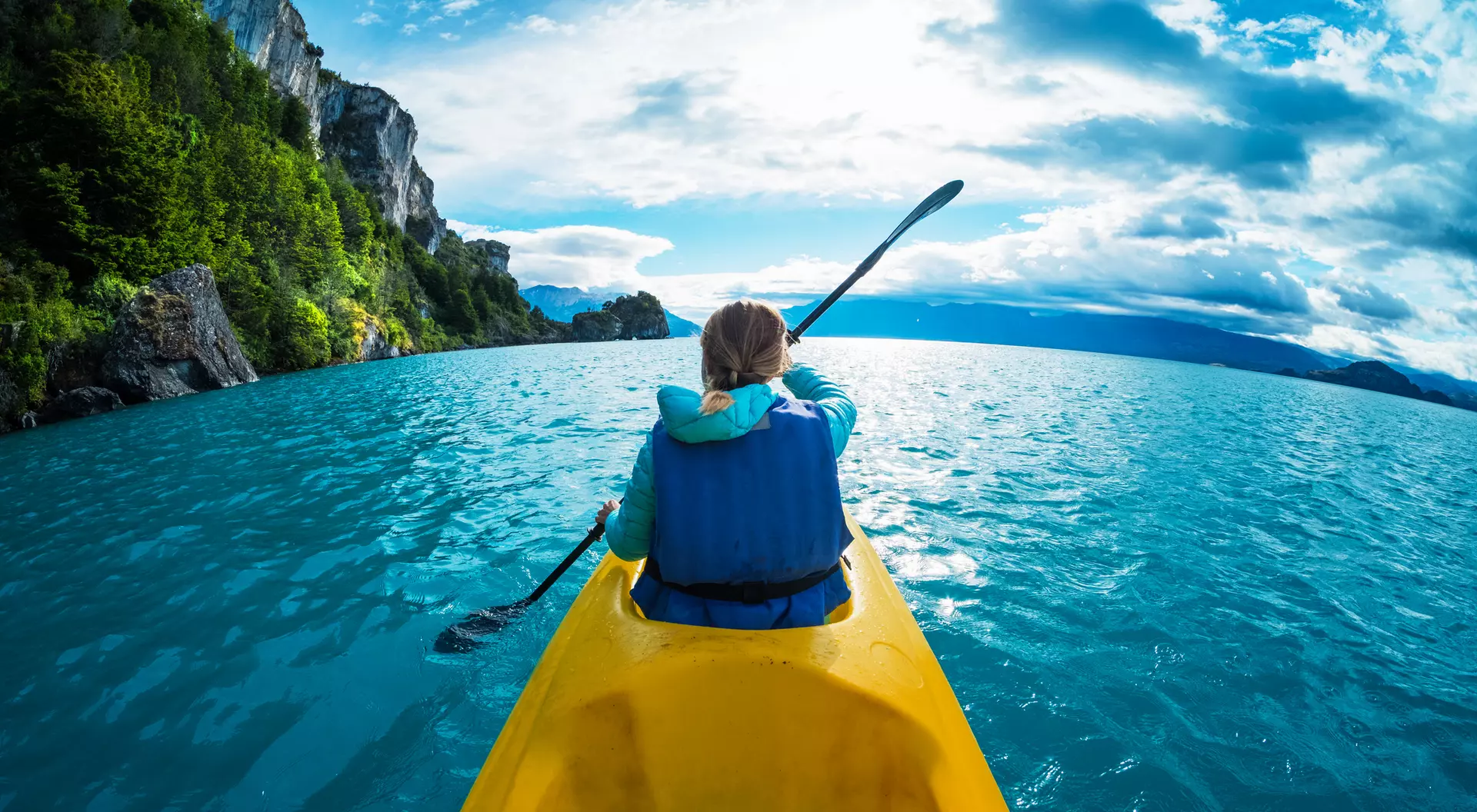 Person kayaking in vibrant blue waters with mountains and cloudy sky in the background.