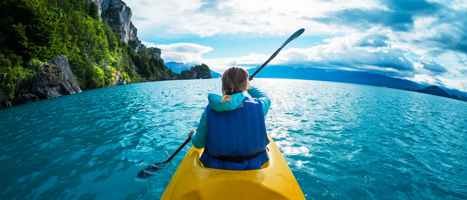 Person kayaking in vibrant blue waters with mountains and cloudy sky in the background.