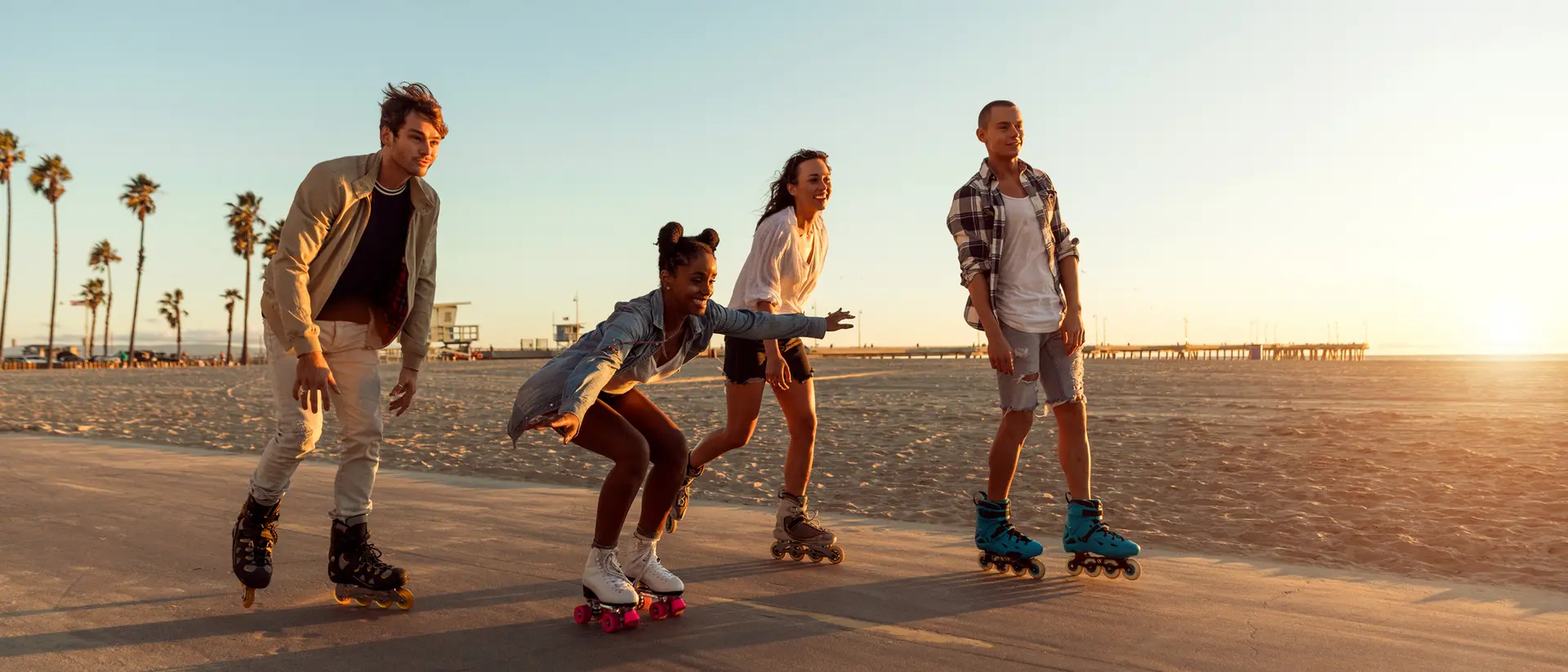 Four friends roller skating along a beachside path during sunset, with palm trees in the background.