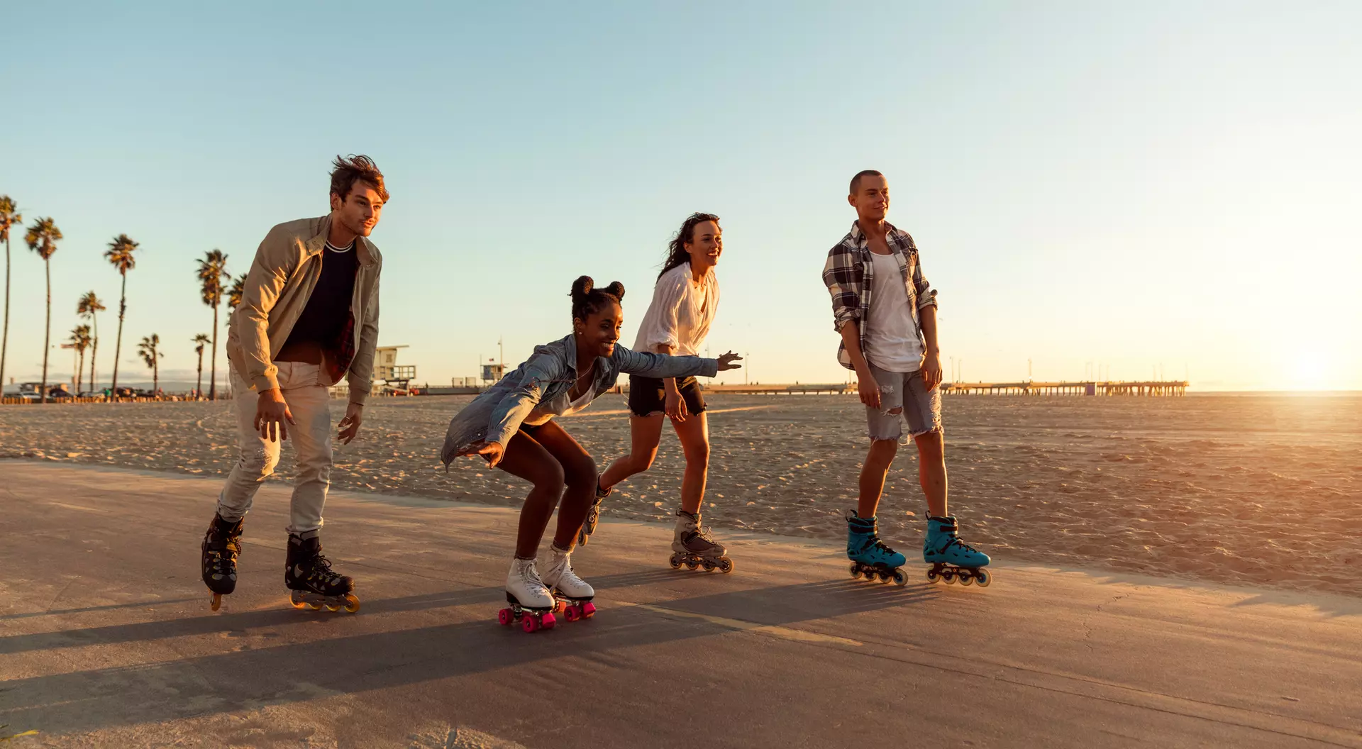 Four friends roller skating along a beachside path during sunset, with palm trees in the background.