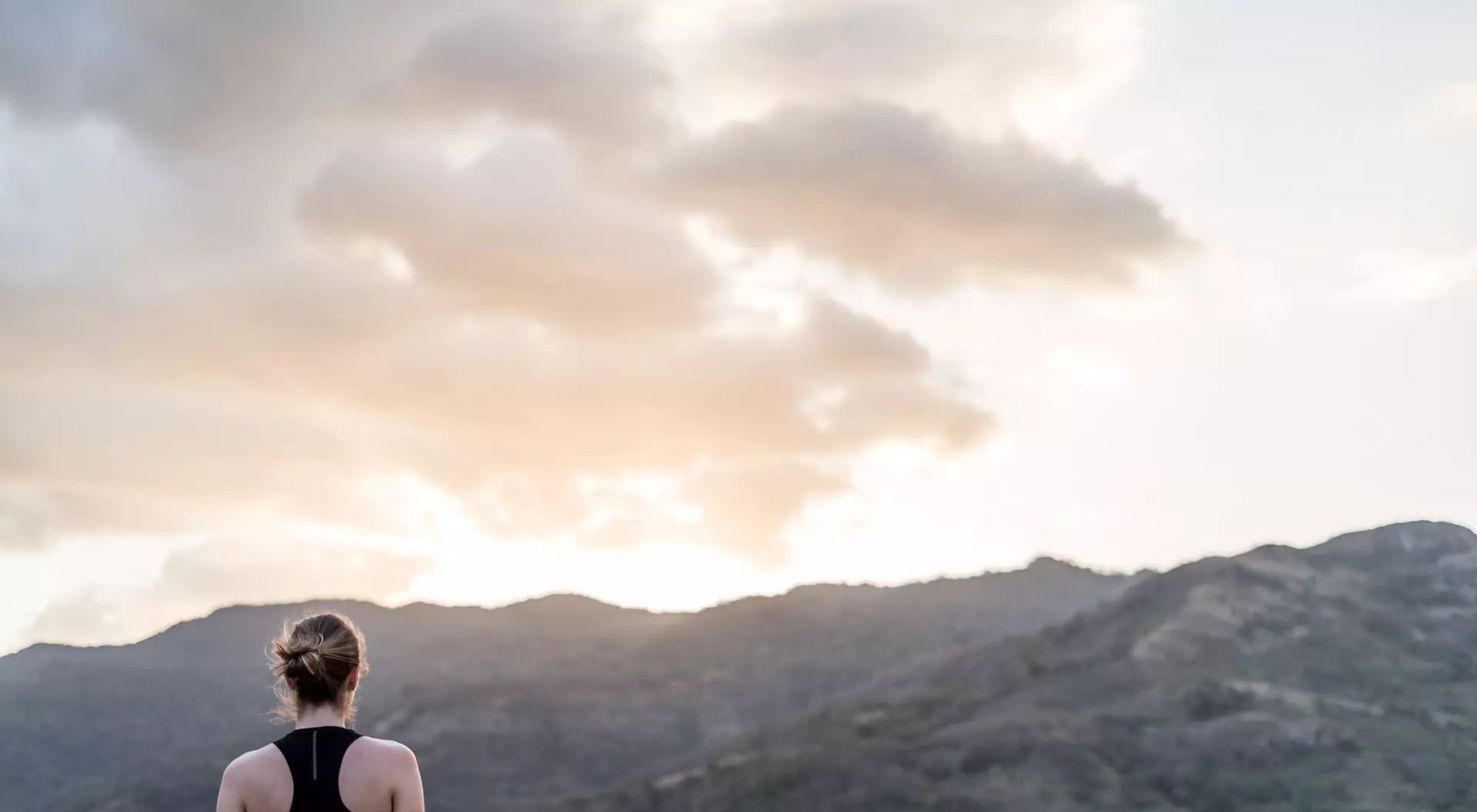 A person stands facing distant mountains and a sunset, with soft clouds in the sky.