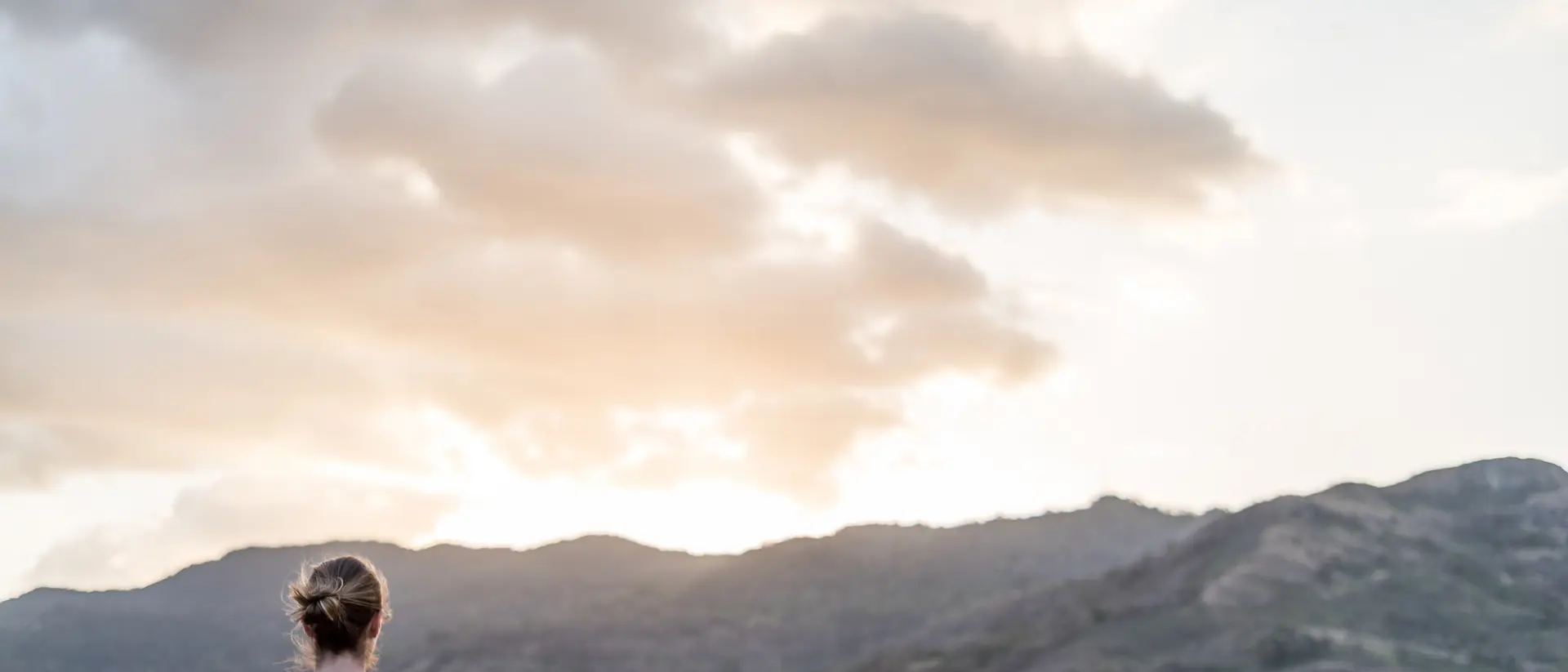 A person stands facing distant mountains and a sunset, with soft clouds in the sky.