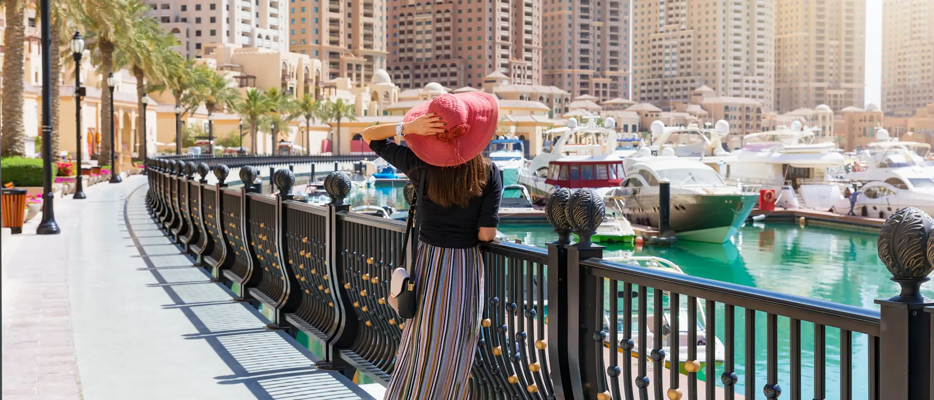 A person in a wide-brimmed hat strolls along a waterfront promenade, with boats and tall buildings in the background.