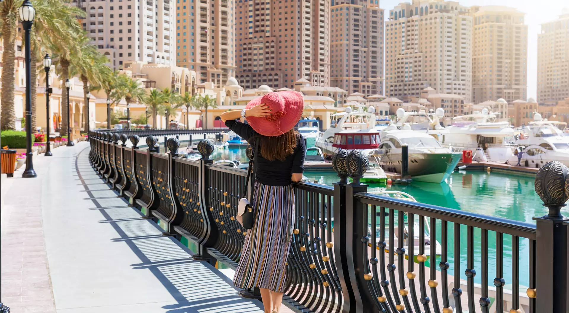 A person in a wide-brimmed hat strolls along a waterfront promenade, with boats and tall buildings in the background.