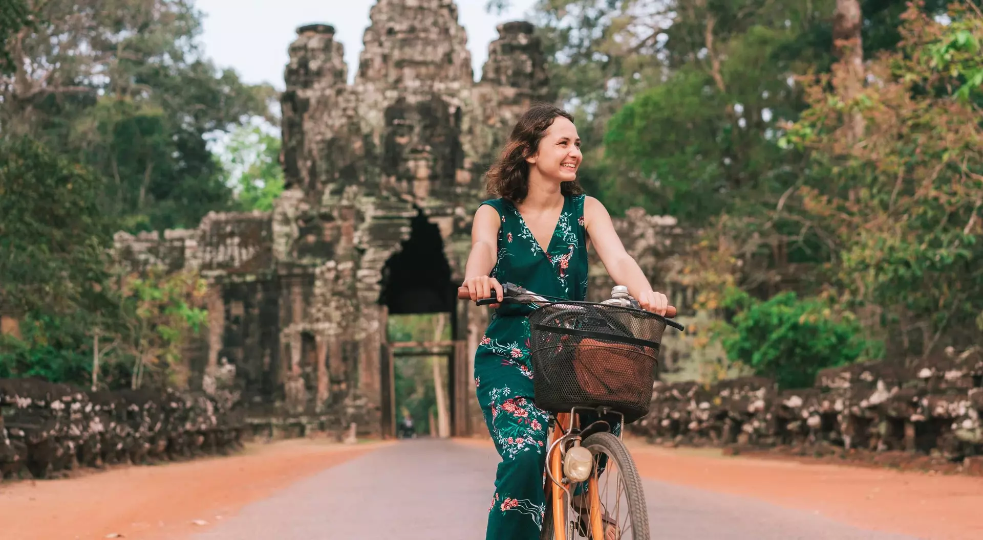 A woman rides a bicycle on a path near a historic stone temple surrounded by lush greenery.