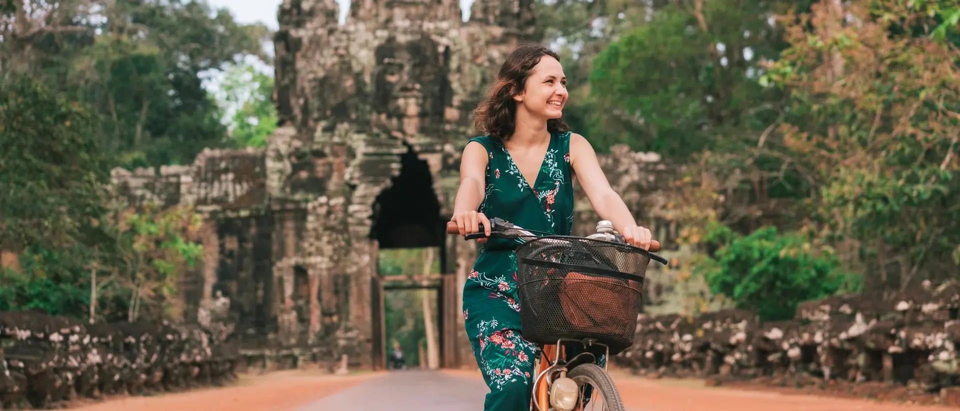 A woman rides a bicycle on a path near a historic stone temple surrounded by lush greenery.