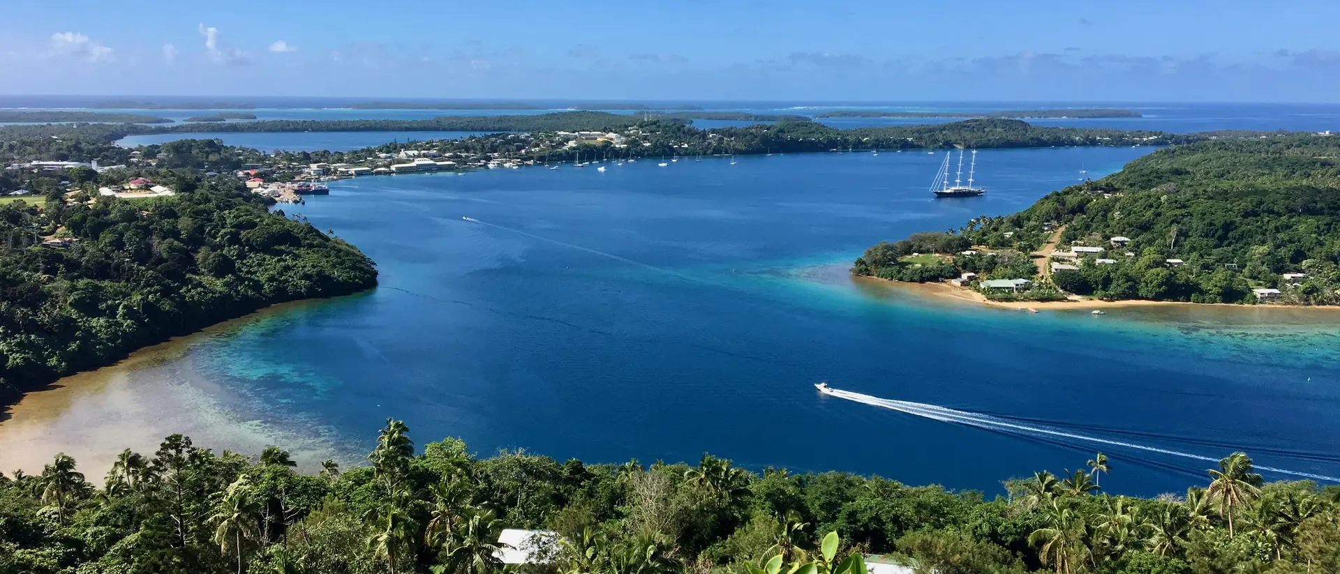 A scenic view of a bay with clear blue water, surrounded by greenery and small boats, under a bright blue sky.