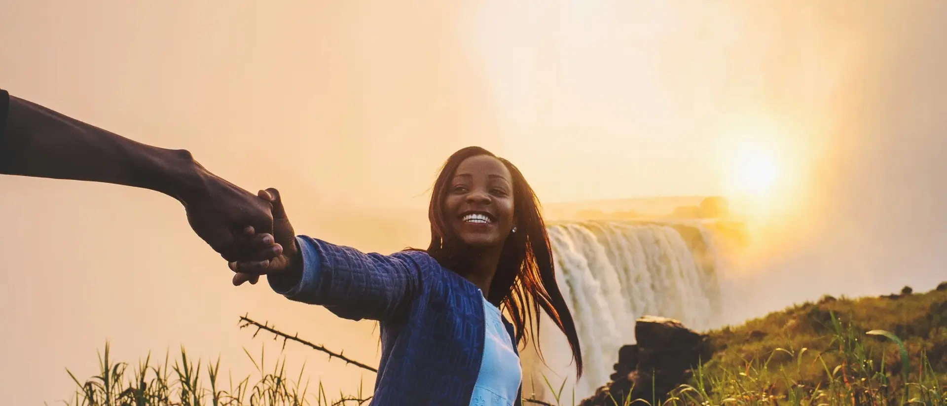 A woman joyfully holds hands with a person, standing near a waterfall at sunset amidst lush greenery.