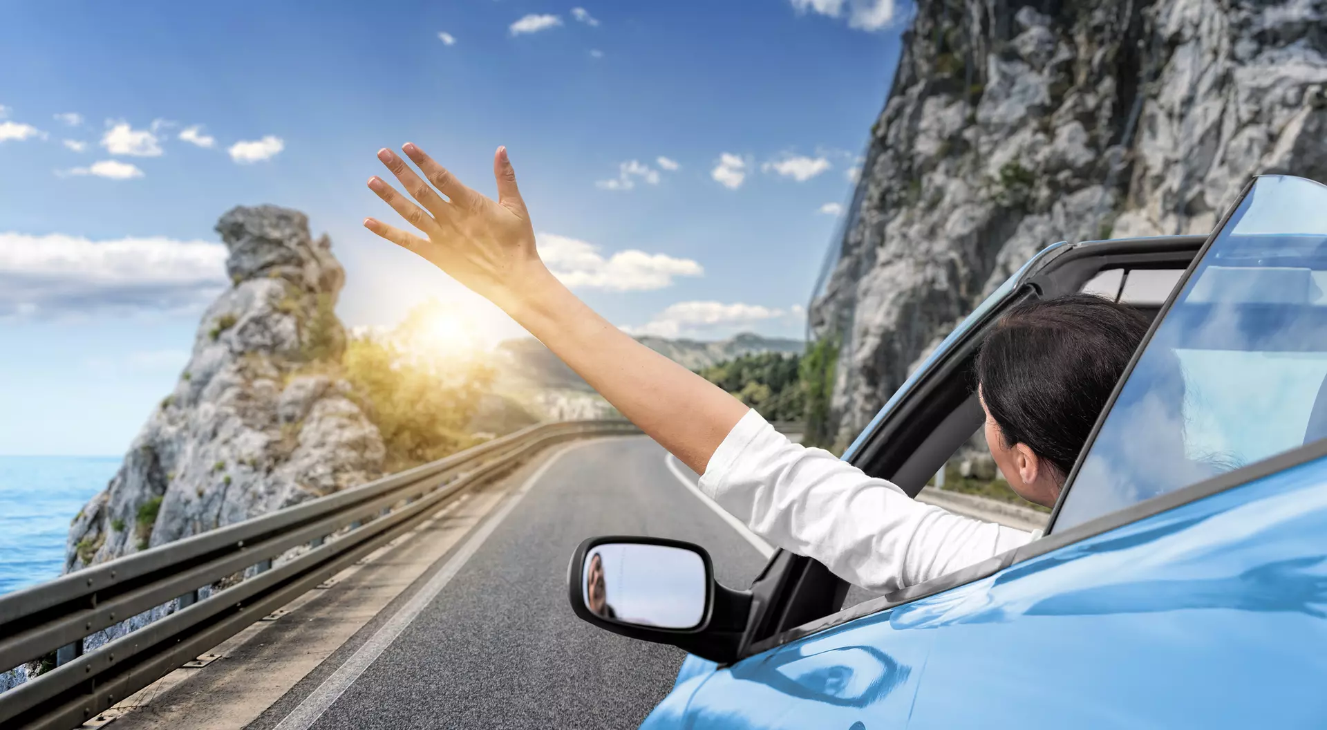 A person in a blue car waves their hand out of the window, driving along a scenic coastal road with cliffs and sunlight.