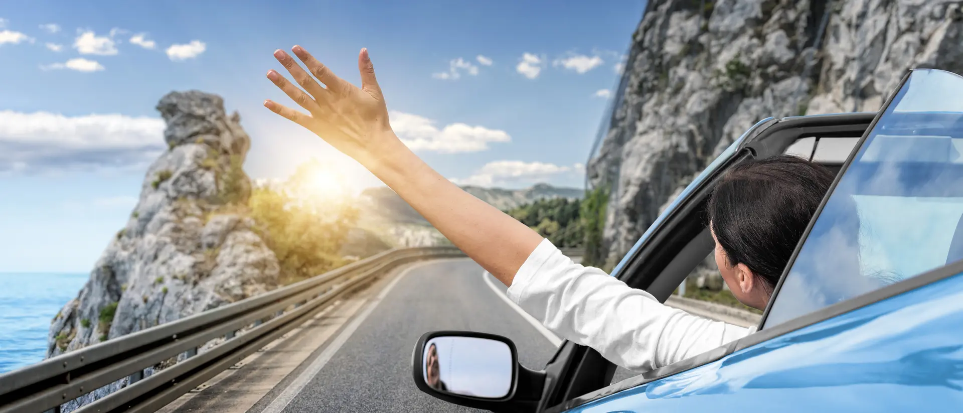 A person in a blue car waves their hand out of the window, driving along a scenic coastal road with cliffs and sunlight.