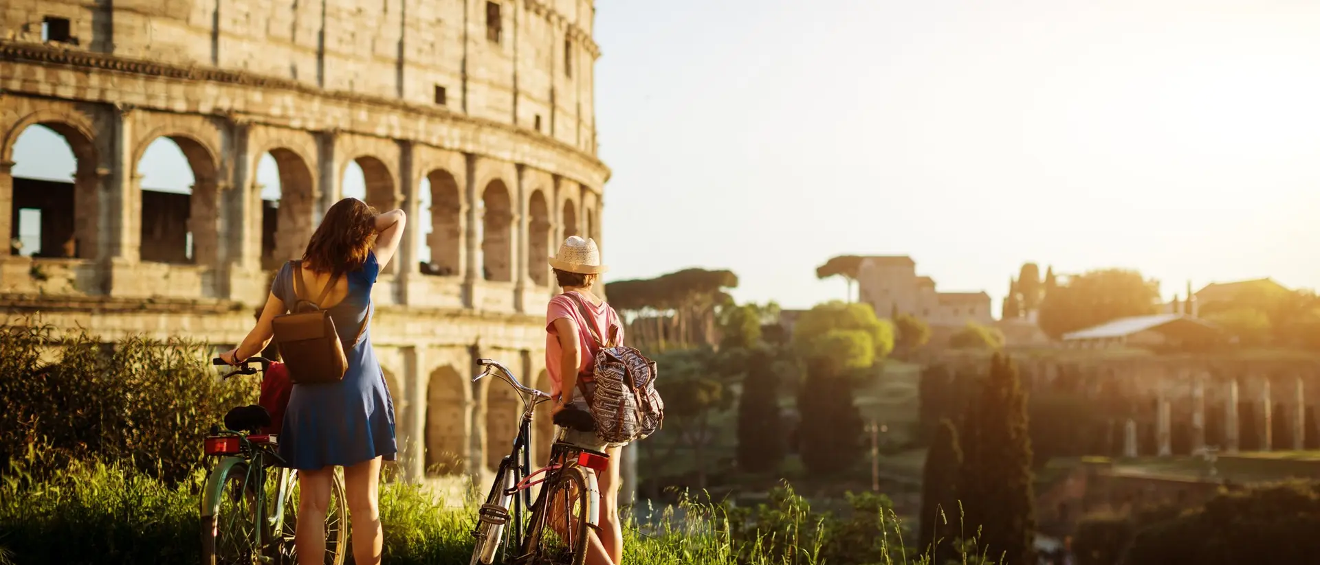 Two friends stand with bicycles, admiring the Colosseum in Rome at sunset, surrounded by greenery.