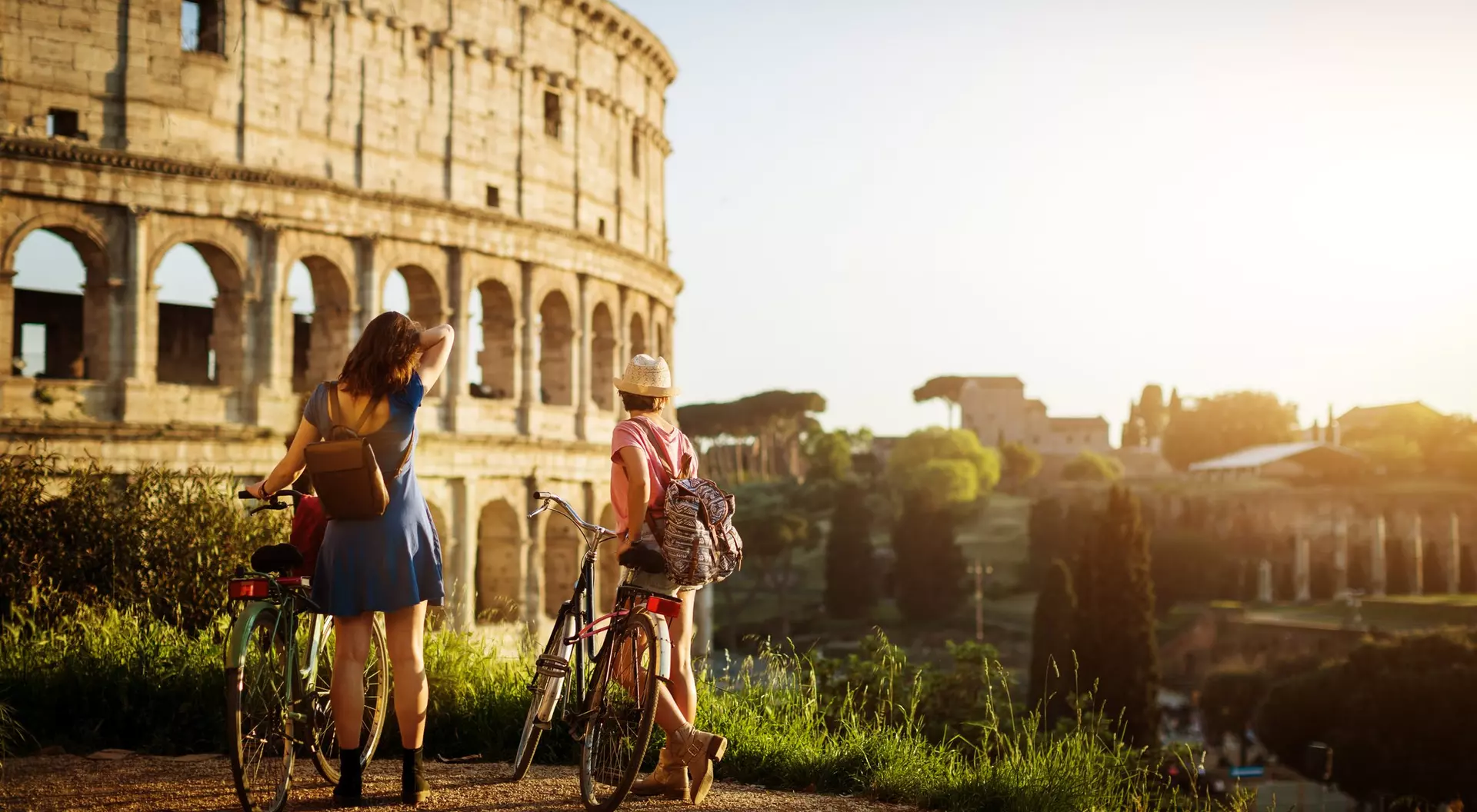 Two friends stand with bicycles, admiring the Colosseum in Rome at sunset, surrounded by greenery.