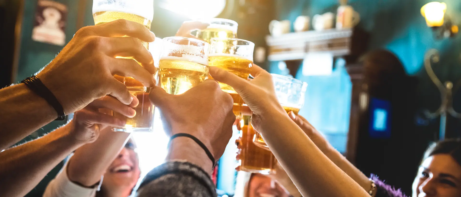 Group of people raising glasses of beer in a toast, smiling, inside a lively pub with colorful decor.