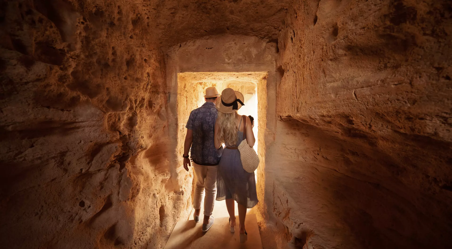 A couple walks hand-in-hand through a narrow, lit stone passageway, surrounded by textured walls.