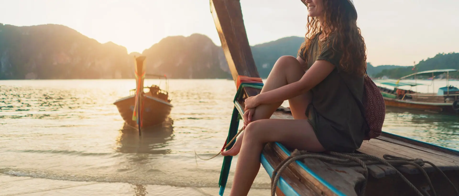 A young woman in a wide-brimmed hat sits on the edge of a boat, enjoying a sunset over the water.