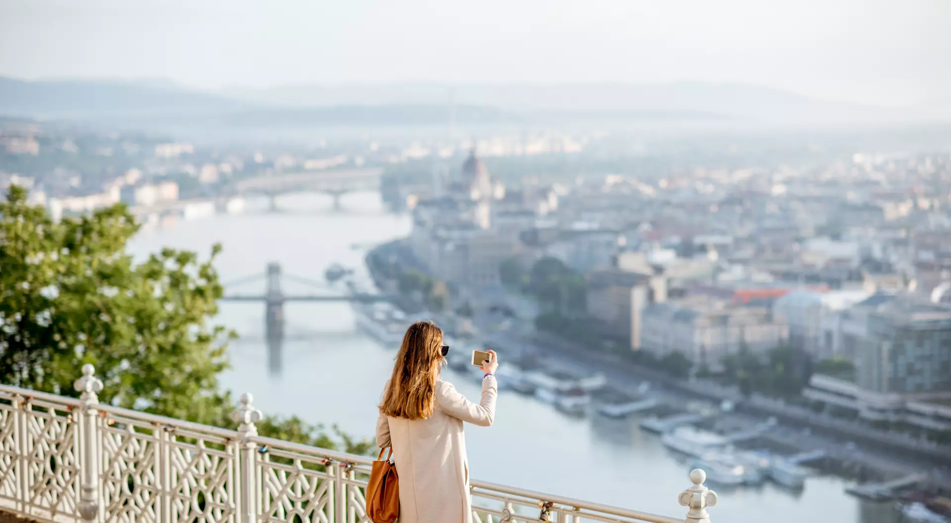 A woman in a cream coat enjoys a scenic view of a city and river from an elevated lookout point.