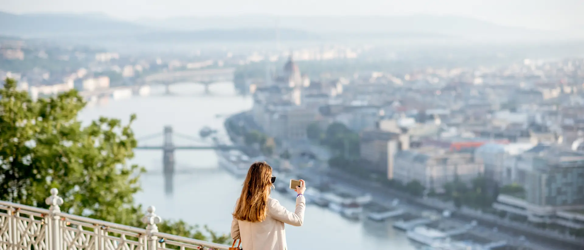 A woman in a cream coat enjoys a scenic view of a city and river from an elevated lookout point.