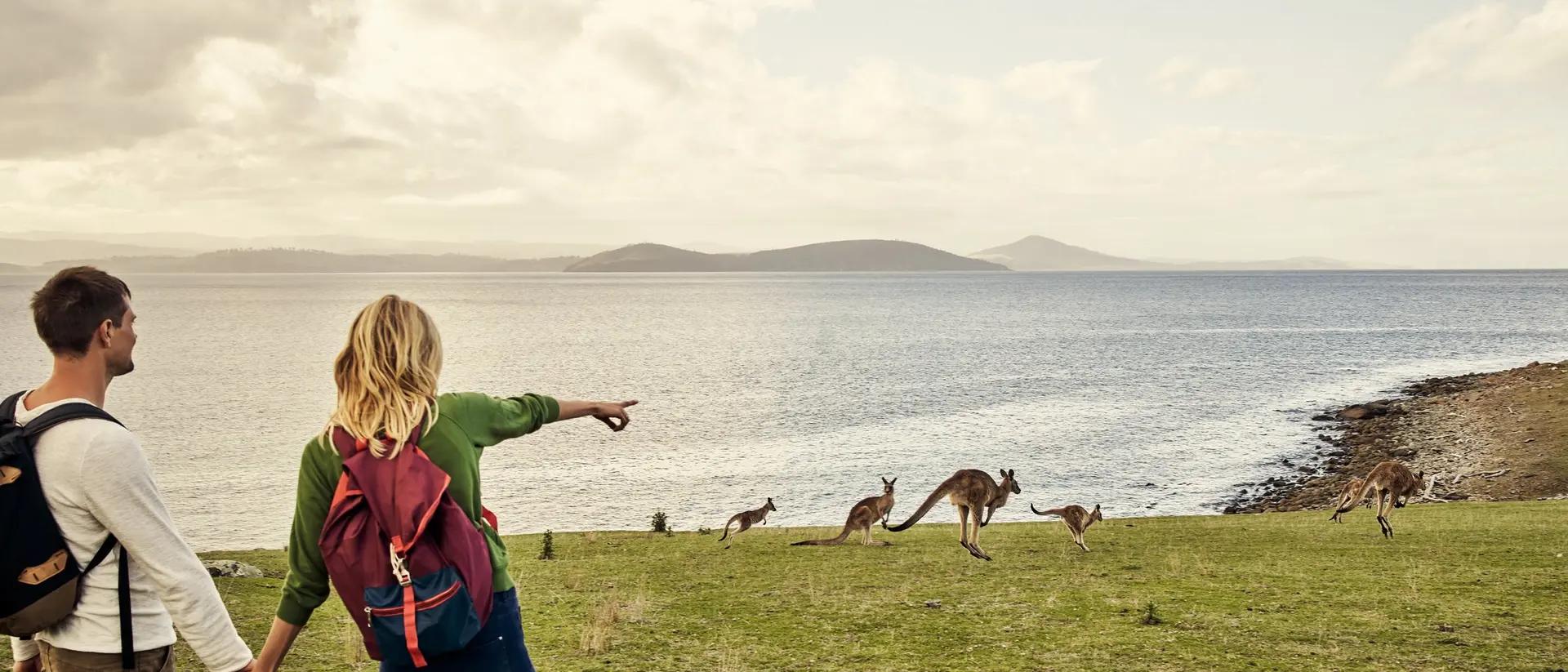A couple stands by the water, pointing at kangaroos on the shore under a cloudy sky.