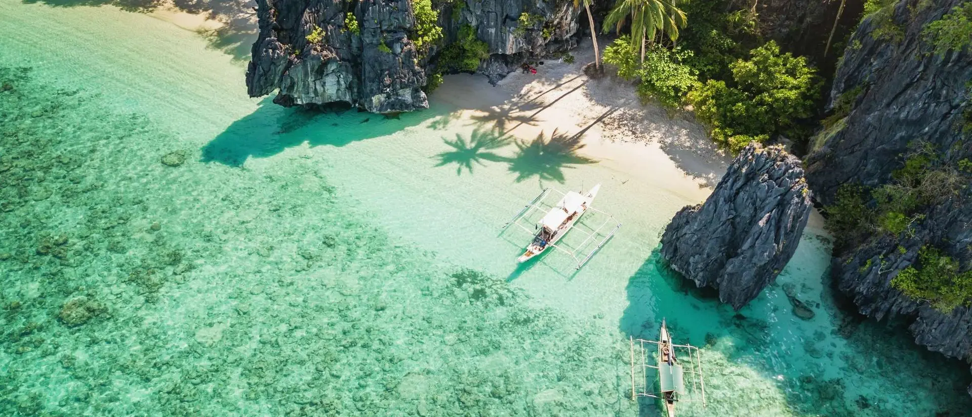 Aerial view of a clear turquoise sea, sandy beach, lush greenery, and two boats near rocky cliffs.