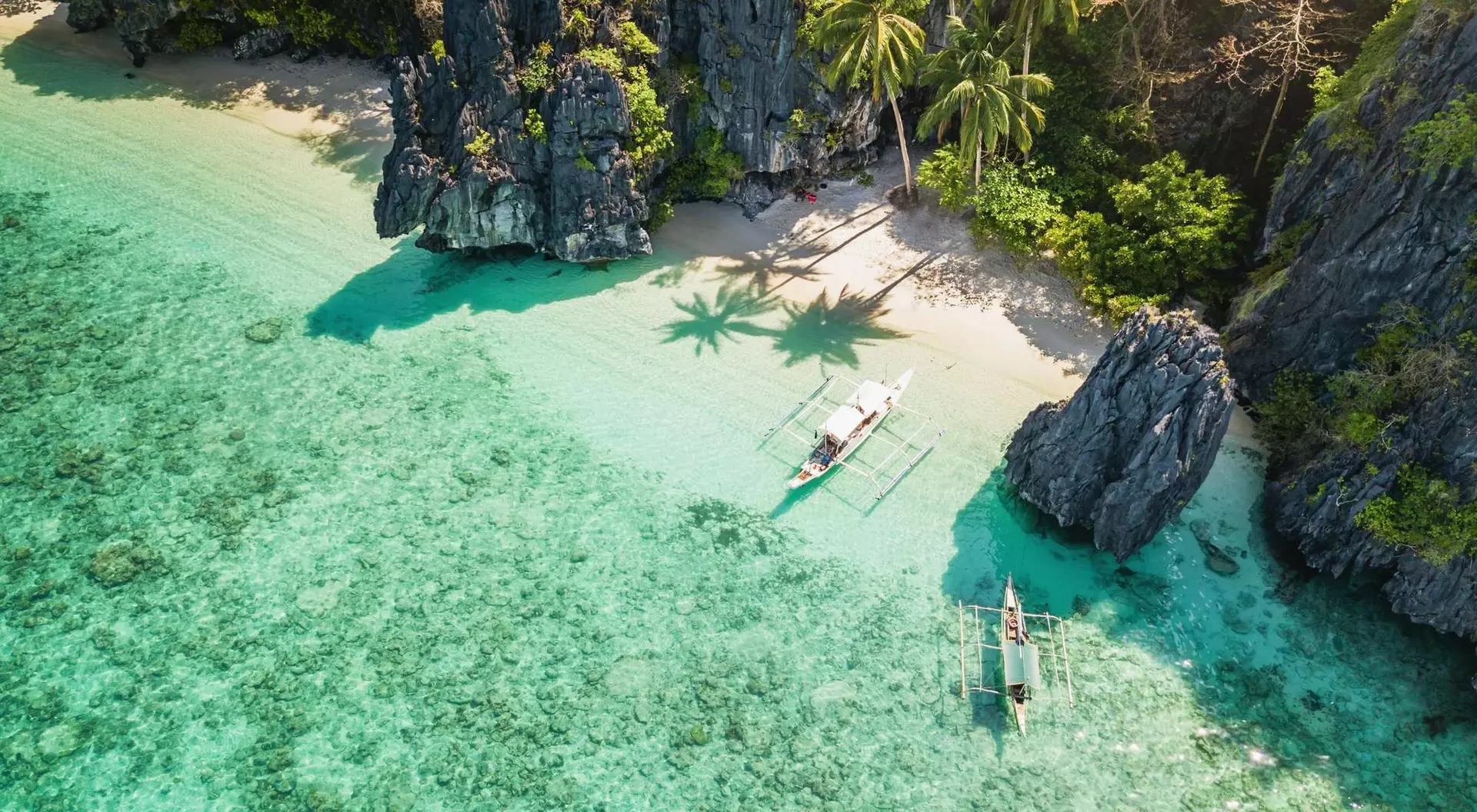 Aerial view of a clear turquoise sea, sandy beach, lush greenery, and two boats near rocky cliffs.