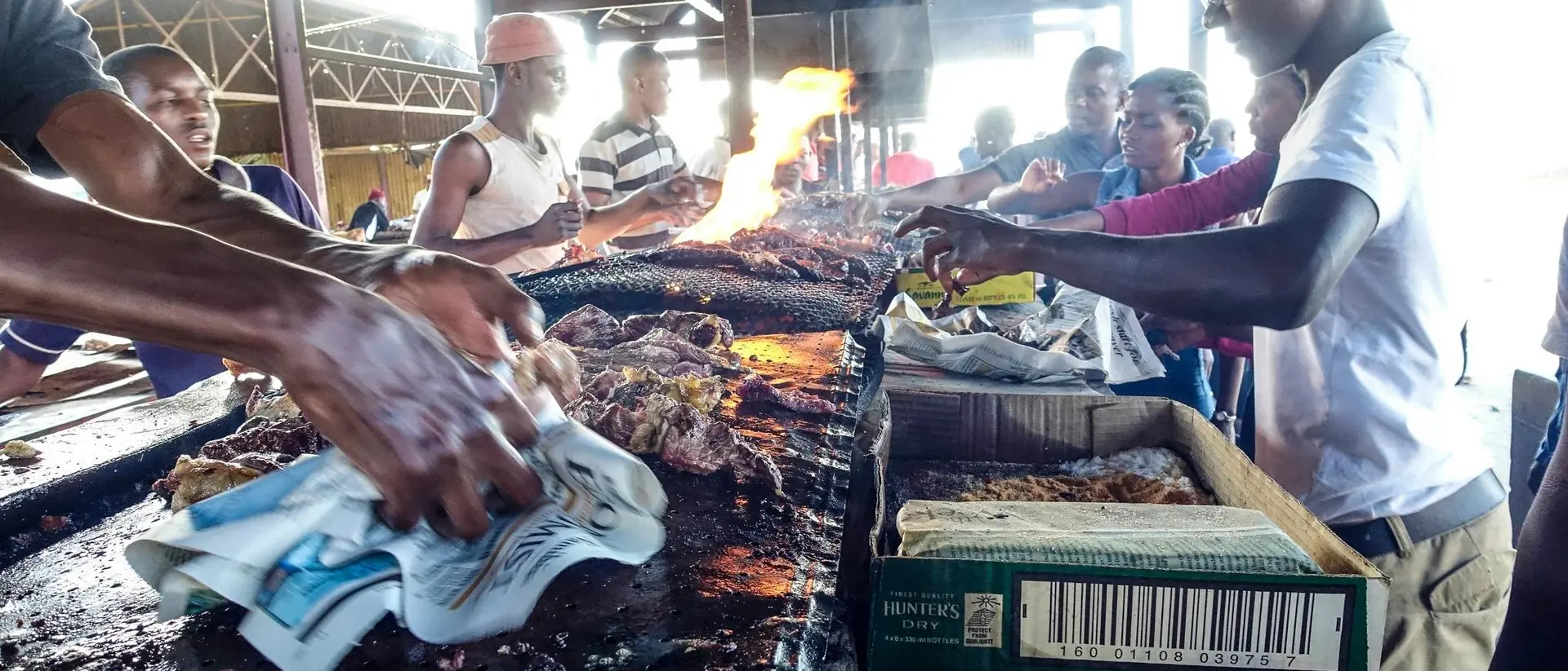 A bustling market scene with people grilling food over open flames, surrounded by vendors and onlookers.
