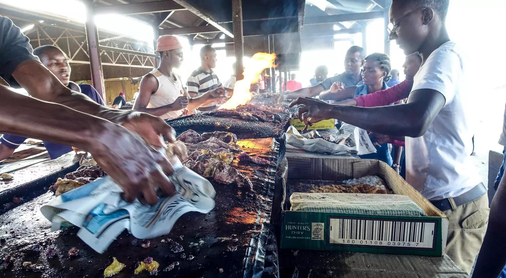 A bustling market scene with people grilling food over open flames, surrounded by vendors and onlookers.