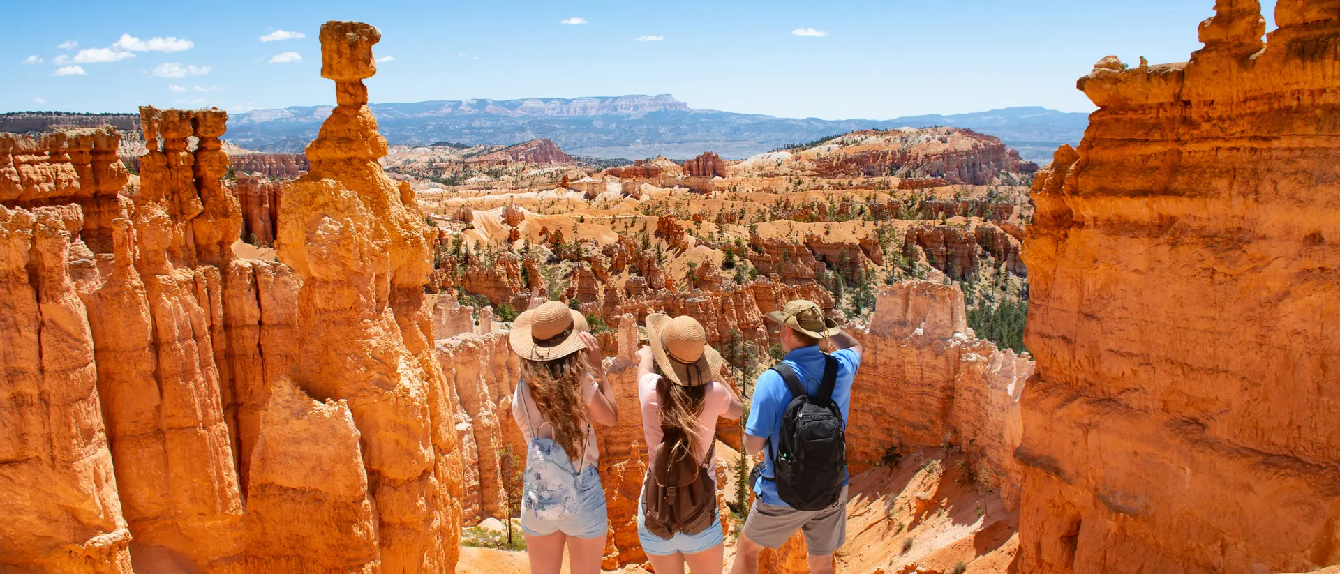Three travelers stand at the edge of a canyon, gazing at orange rock formations under a clear blue sky.